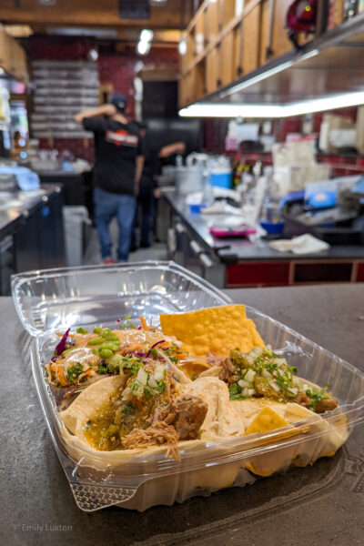 Clear plastic takeaway food container on a bar in a restaurant with three tacos inside loaded with meat and guacamole. 