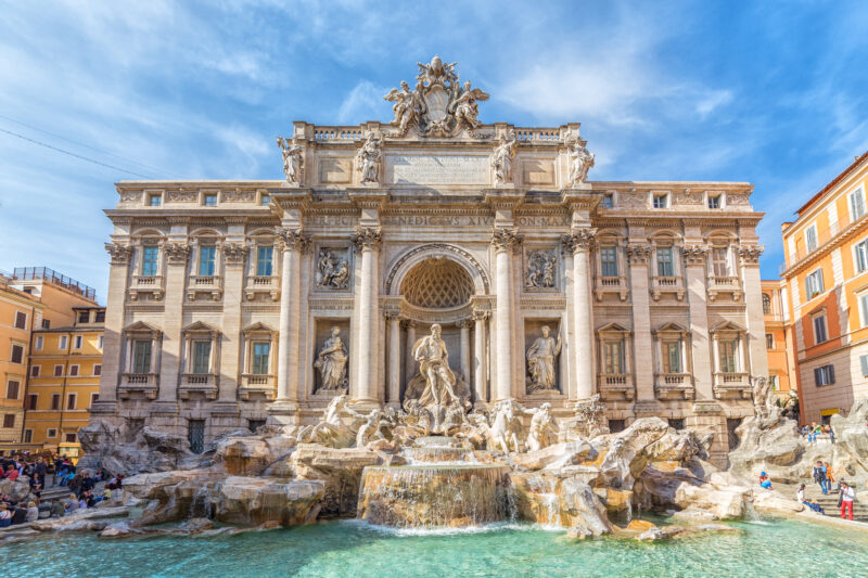 Trevi Fountain in Rome, Italy, a large bright blue pool with a crowd of stone carvings and statues behind it and a large cream coloured building behind with columns and more statues in arched nooks 