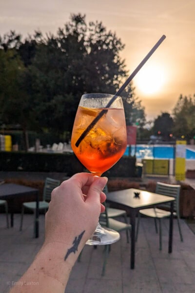 hand holding a large glass of aperol spritz, a bright orange drink with large chunks of ice and a black straw. There are tables and a swimming pool out of focus behind and the sun is setting in a golden sky. The hand has a small black tattoo of a pterodactyle on the wrist. 