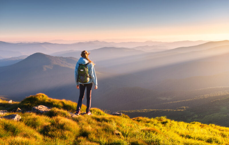 Beautiful mountains in fog and standing young woman with backpack on the peak at sunset in summer. 