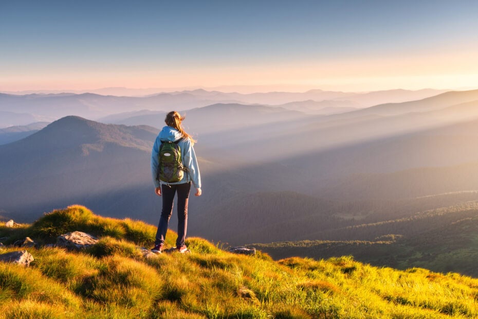 Beautiful mountains in fog and standing young woman with backpack on the peak at sunset in summer.