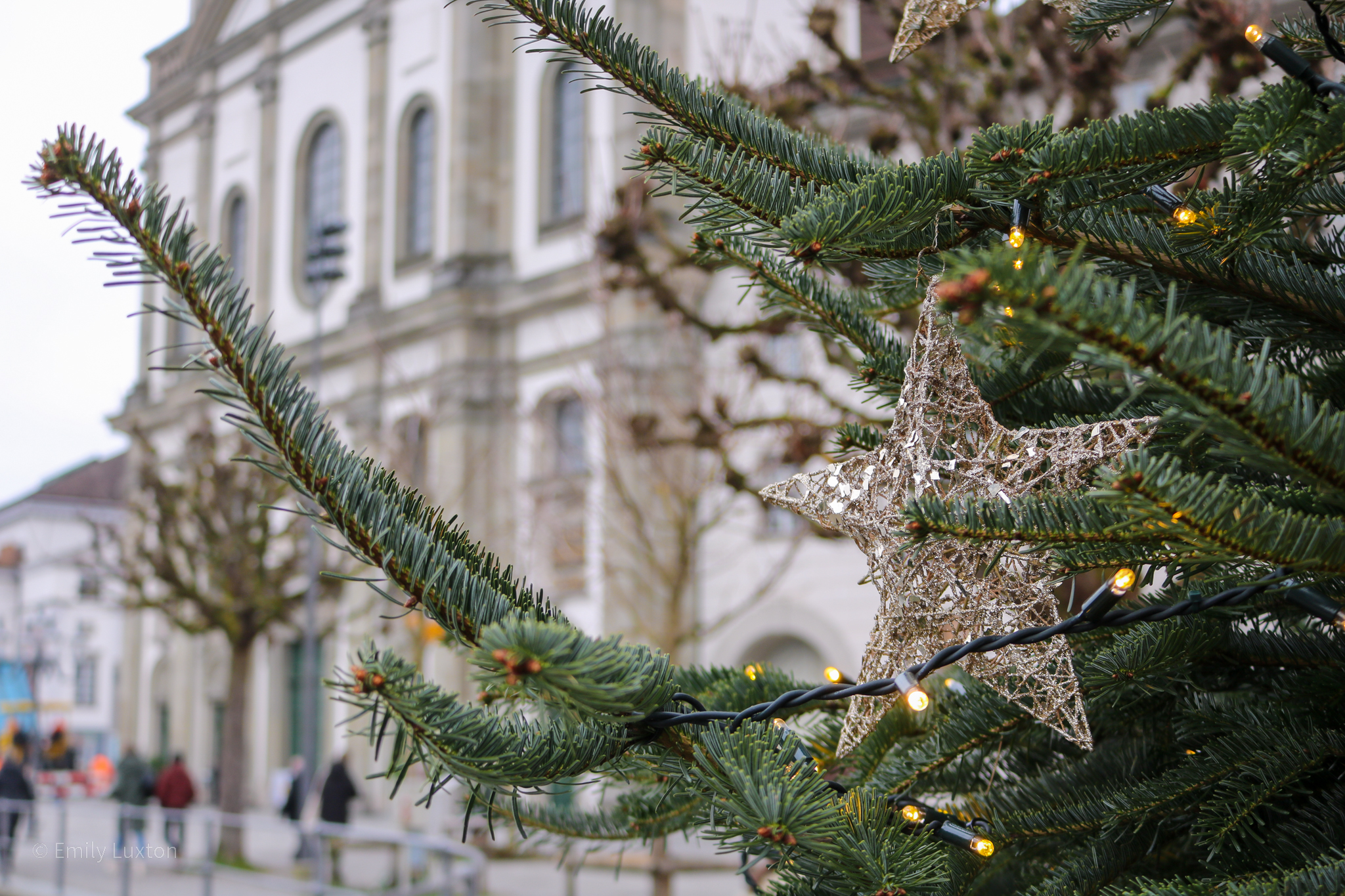 Close up of the side of a christmas tree with a wooden star hanging from a branch. There is a white church behind but it's out of focus.