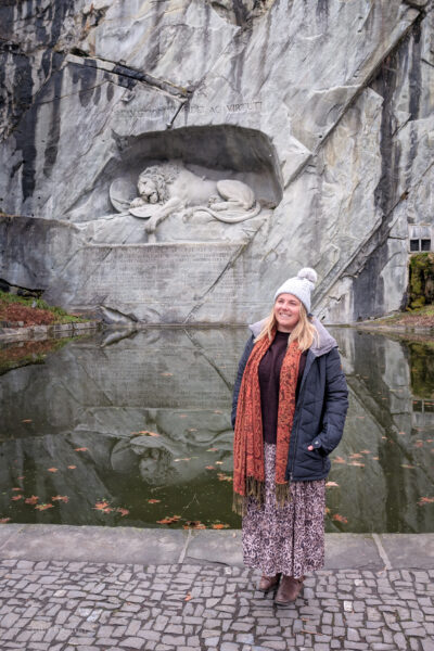 Emily standing in front of a small pond with a sheer grey rock face behind. There is a statue of a sleeping lion carved into the rock. Emily has her hands in her coat pockets and is wearing a burgundy jumper, an orange scarf, a grey wooly bobble hat, a leopard print skirt, and brown boots.