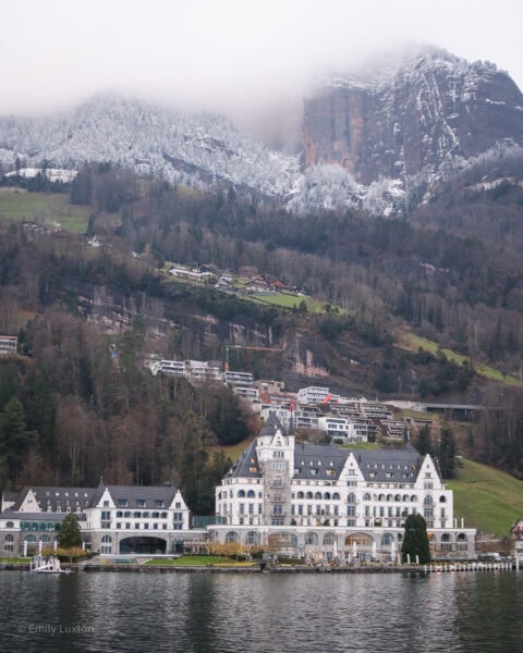 large white manor type building with a grey roof at the foot of a towering mountain with snowy peaks. There is a still grey lake in front.