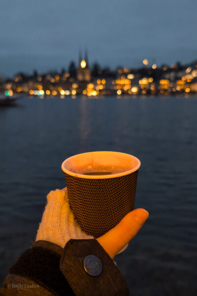 gloved hand holding a paper cup with red wine in front of a dark blue lake at night time with city lights out of focus on the far side of the lake.