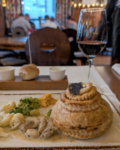 Recangular plate with a puff pastry pie next to some meat and vegetables in a creamy sauce. There is a small glass of red wine next to the plate and the rest of the restaurant is out of focus beyond.