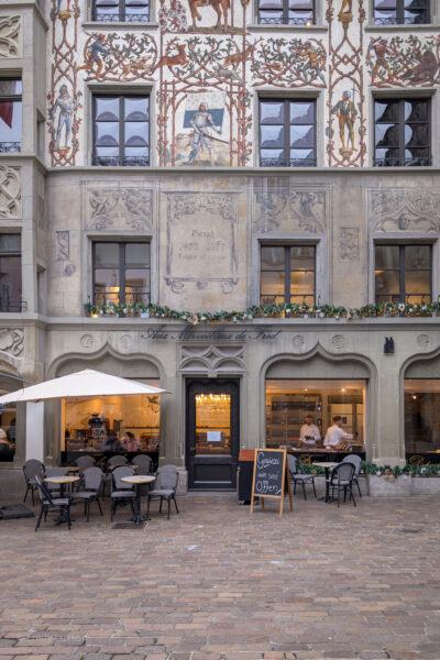 Square in Lucerne Old Town with dark grey flagstone floor. There is an old building with a grey stone facade, the top three floors are painted cream with a colourful mural depicting knights in armour and deer.