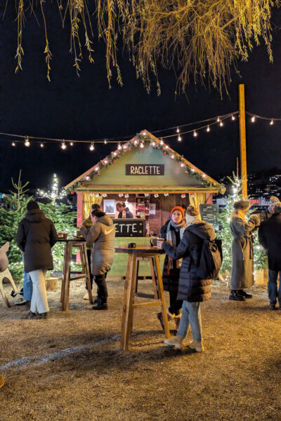 Small wooden hut painted green with a sign on the rood reading "Raclette". The hut is surrounded by small christmas trees with string ligths running overhead. It is night time and several people are standing in front of the hut at tall wooden tables.