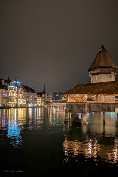 Covered wooden bridge over a river at night with a circular stone water tower behind, both have red tilded roofs. The city lights on the far side of the river are reflected in the river.