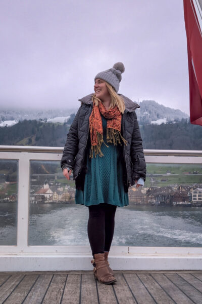 Emily standing on the stern of a boat facing the camera with a view of the lake and snowcapped mountains nehind her and a small town at the edge of the lake. She is wearing a long grey winter coat with a fur lined hood, a dark teal wool dress, black leggings, brown leather boots and a light grey hat. The swiss flag is flying from the back of the boat next to her.