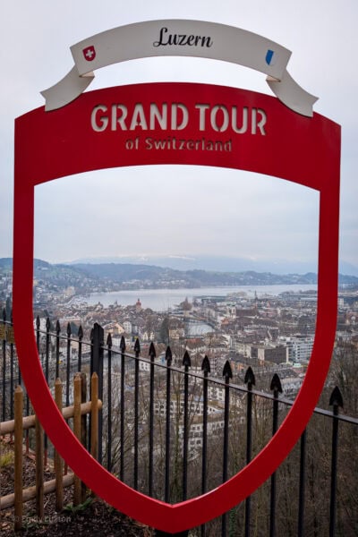 View of Lucerne city centre and the neighbouring lake from a hilltop on a grey overcast day in winter. The view is seen through a red wooden frame with a white sign on top that says Luzern, and white letters below that reading Grand Tour of Switzerland. 