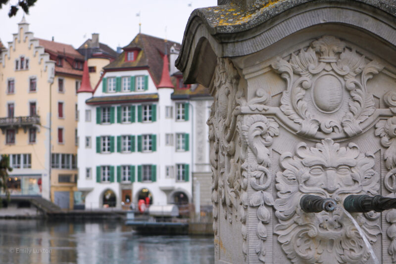 close up of a stone fountain with a carved face of a myhtical figure around the spout, in the background is a river and historic swiss style architecture.