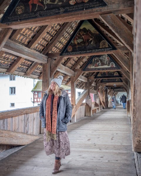 emily walking through a wooden covered bridge with her hands in her pockets. The floor is wooden and there is a triangular wooden roof above, while the sides of the bridge are open with a view of a riverside building beyond. Emily is wearing a grey winter coat, a long leopard print skirt, brown leather boots, a grey wool bobble hat and an orange scarf. 