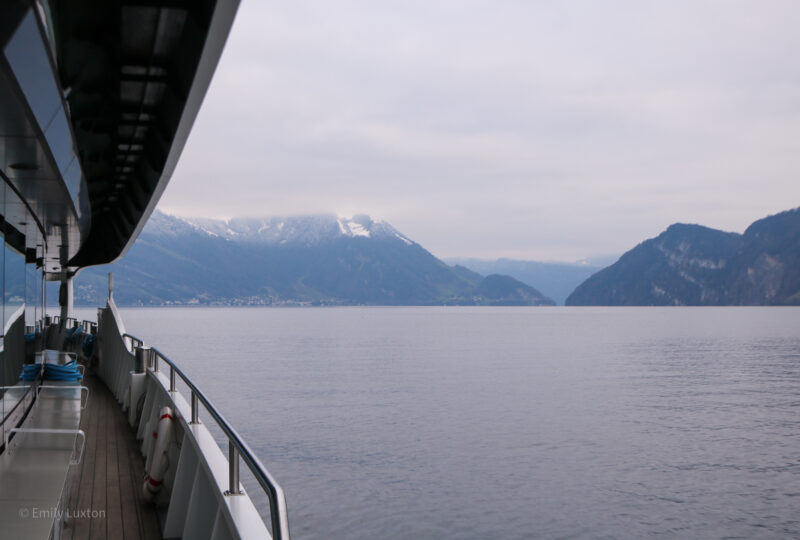 Looking along the side of a boat on a lake with very calm grey water and a mountain range beyond. It is a cold day in winter with grey sky above Lake Lucerne.