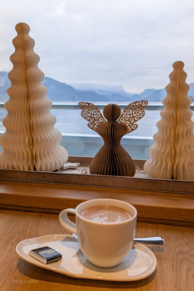 small white mug filled with hot chocolate on a plate on a wooden table next to a window. There are some paper christmas decorations of an angel and two christmas trees on the window ledge. Outside is a view of a lake and mountains. 