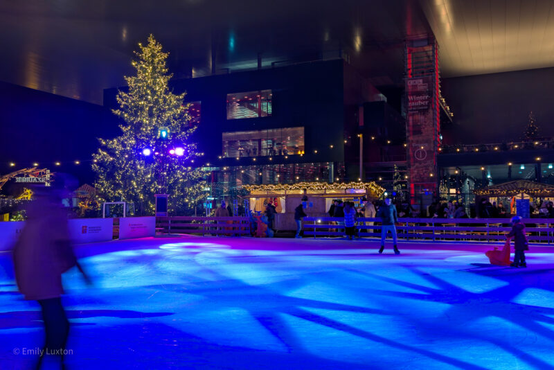 Outdoor ice rink at night lit up in blue with star-shaped patterns on the floor and blurry human figures skating by. There is a large Christmas tree covered in yellow lights behind. 