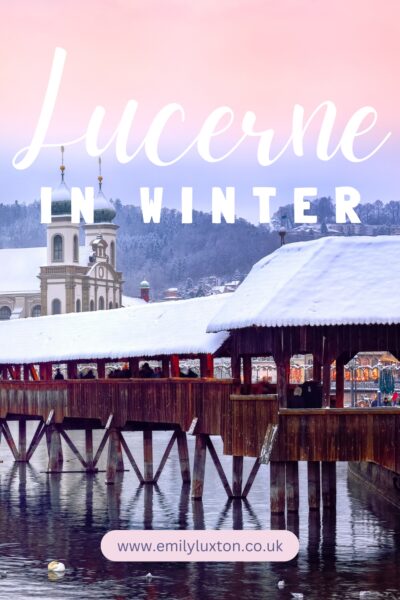 Lucerne in Winter: written in white text on top of an image of a covered wooden bridge covered with heavy snow an dreflected in the still grey water of the river. There is a historic white jesuit church behind with green doems on top of the two towers and pink sunset sky overhead. 