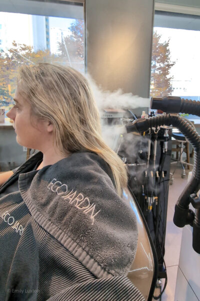 Emily is seated in a salon undergoing a hair treatment with a grey towel labeled "ECOJARDIN" draped over her shoulders. A machine emits steam directed at Emily's hair as part of the 15 Step Korean scalp treatment in Seoul. The background features salon equipment and a window with a view of buildings and trees outside.