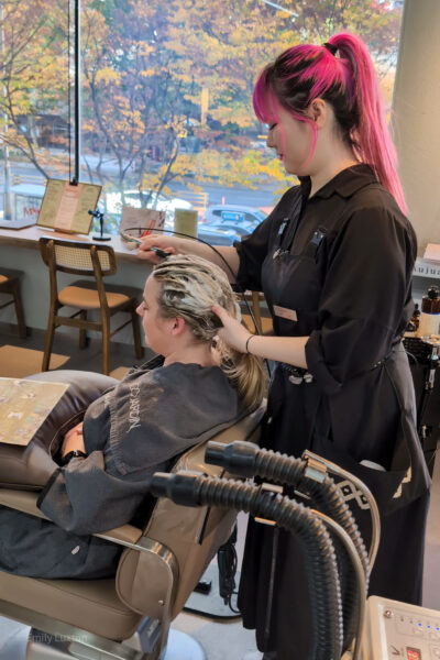 Scene inside a hair salon where a Korean woman with pink hair, wearing a black apron, applies shampoo to Emily's hair. Emily is seated in a salon chair, covered with a grey salon cape. The salon has large windows with a view of trees with autumn foliage outside, and chairs and a counter with various items in the background.