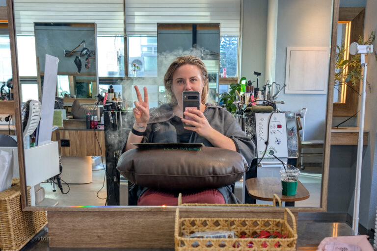 Emily sits in a salon chair, facing a mirror, with their her obscured by her phone which she holds in one hand, making a peace sign with the other. A brown cushion is on her lap. The background includes various salon equipment like hair products, brushes, and a machine with buttons and dials. A drink is on a small table to the right. Emily is wearing a grey salon robe.