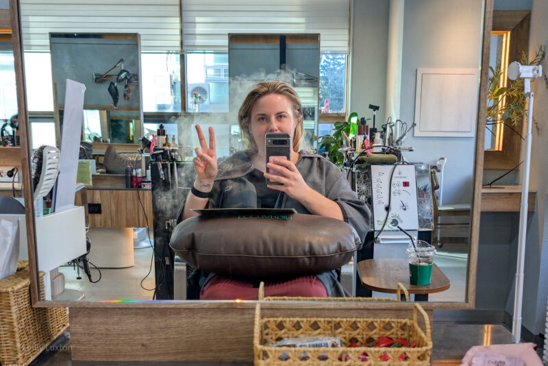 Emily sits in a salon chair, facing a mirror, with their her obscured by her phone which she holds in one hand, making a peace sign with the other. A brown cushion is on her lap. The background includes various salon equipment like hair products, brushes, and a machine with buttons and dials. A drink is on a small table to the right. Emily is wearing a grey salon  robe and is getting the South Korean 15 Step Scalp treatment at Eco Jardin in Seoul. 