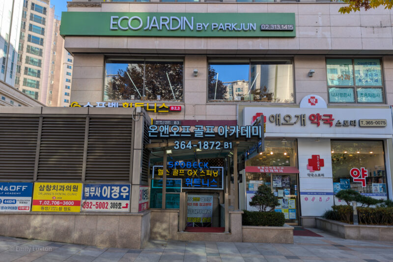 Exterior view of a commercial building with multiple businesses. Signage includes "ECO JARDIN BY PARKJUN" on the upper floor, a pharmacy "애오개 약국 스토리" on the ground floor, "온앤오프 골프 아카데미" (On and Off Golf Academy) in the center, "삼창치과의원" (Dr. Jang's Dental Clinic) on the left, and "법무사 이경우" (Attorney Lee Kyung Woo) also on the left. Various contact numbers are displayed for each business.