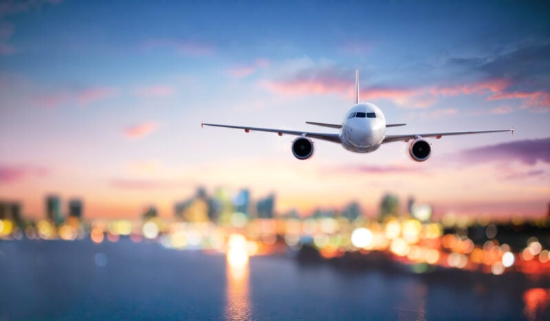 An airplane flying towards the viewer with a city skyline in the background, under a gradient sky of blue and pink hues.