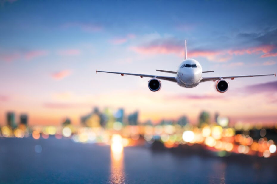 An airplane flying towards the viewer with a city skyline in the background, under a gradient sky of blue and pink hues.