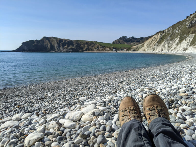 View from a white pebble beach showing brown hiking boots and a calm blue sea with cliffs in the background.