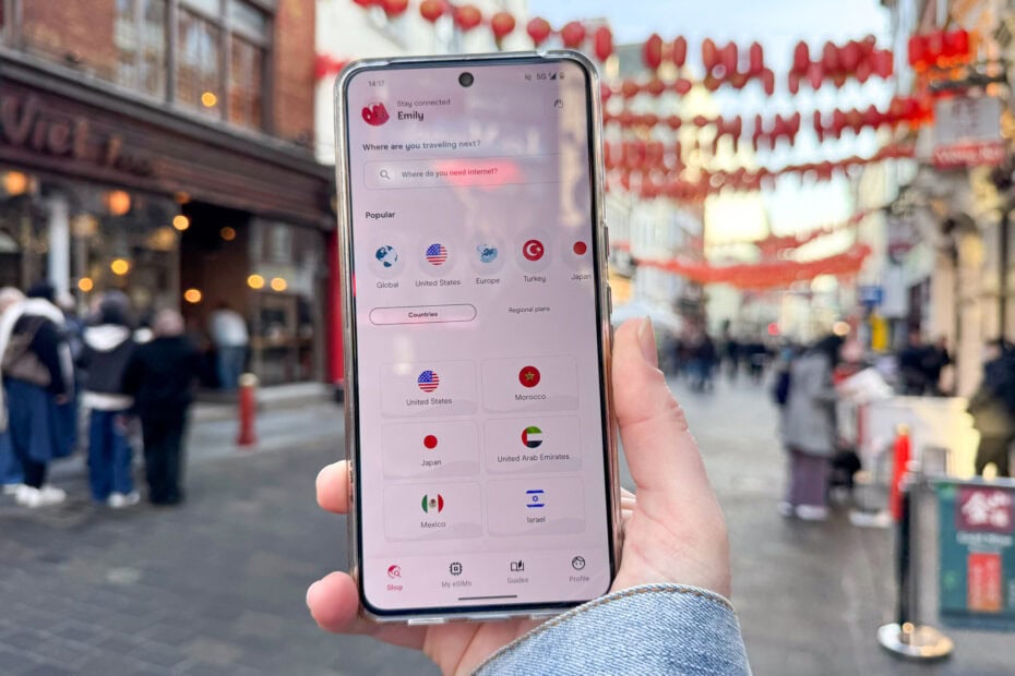 Hand holding a smartphone while standing on a street in Chinatown decorated with red lanterns. The smartphone screen displays an app with the title "Stay connected" and Emily's name at the top. The app includes a search bar with the prompt "Where are you traveling next?" and a highlighted option "Where do you need internet?" Holafly eSIM Review