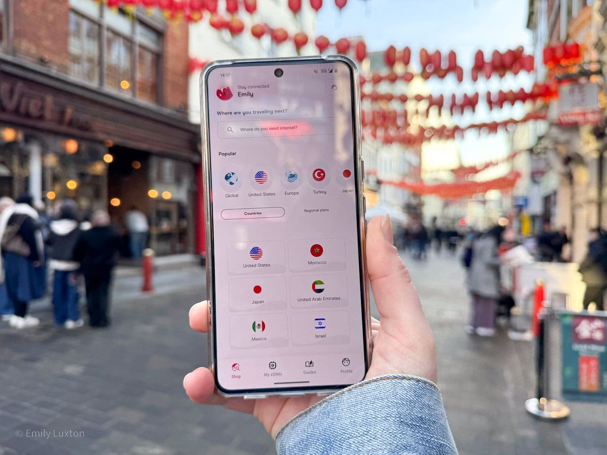 Hand holding a smartphone while standing on a street in Chinatown decorated with red lanterns. The smartphone screen displays an app with the title "Stay connected" and Emily's name at the top. The app includes a search bar with the prompt "Where are you traveling next?" and a highlighted option "Where do you need internet?" Holafly eSIM Review