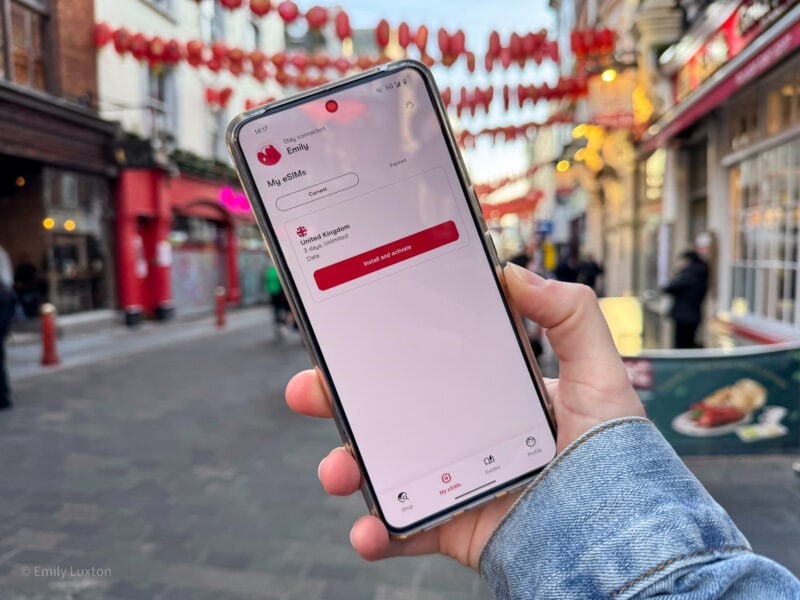 Hand holding a smartphone while standing on a street in Chinatown decorated with red lanterns. The smartphone screen displays an app with Emily's name at the top and the title My eSIMs.