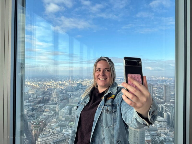 Emily is taking a selfie in front of a large window with a panoramic view of a cityscape. She is wearing a denim jacket and holding a smartphone with a pink pop socket. The background shows a clear blue sky with some clouds and a the London cityscape below.