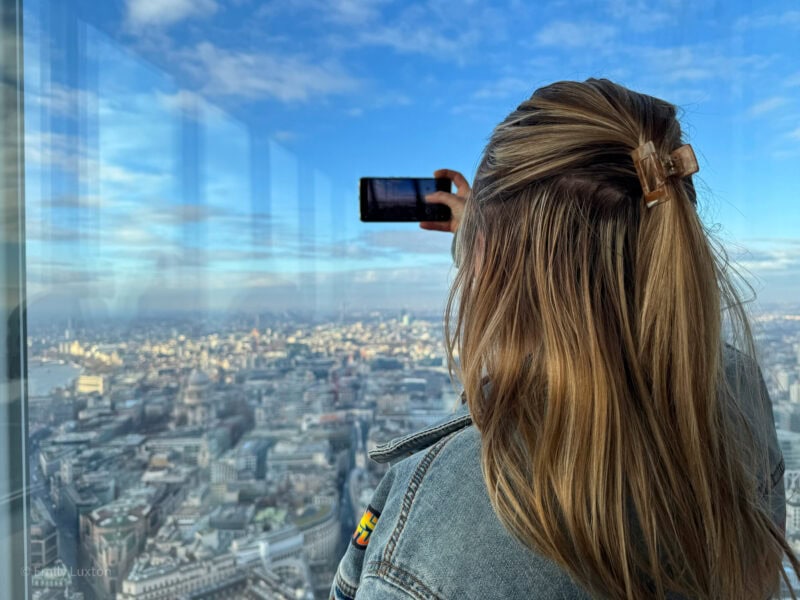 Emily, with long blonde hair held by a clip, is taking a photo with a smartphone from a high vantage point. The view through the glass window shows the London cityscape under a partly cloudy blue sky. The city below is detailed with various buildings and landmarks. Holafly eSIM Review