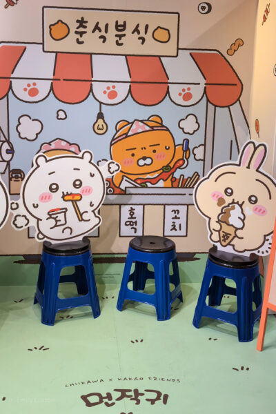 Three blue stools with black seats are placed in front of a cartoon backdrop depicting a food stall with cute characters and Korean text.