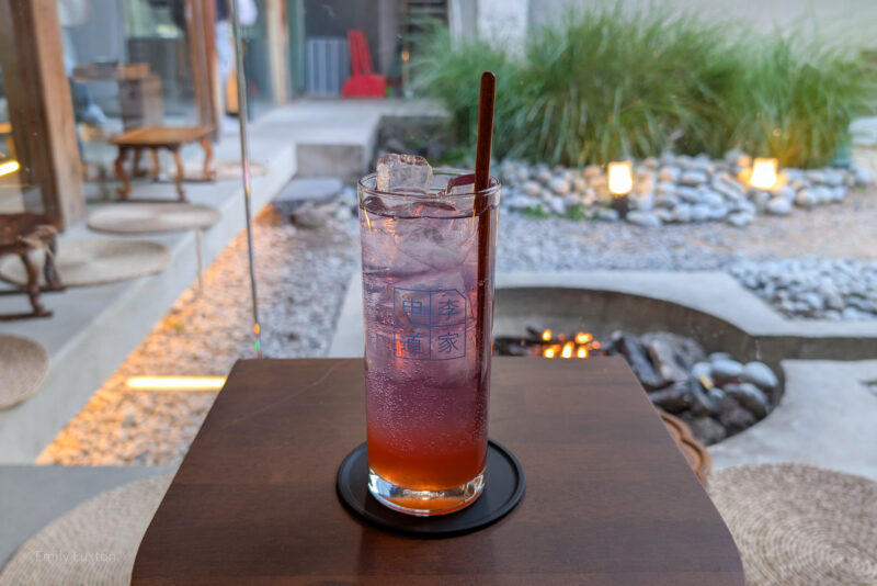 A tall glass filled with a purple and orange gradient drink, topped with ice cubes and a brown straw, sits on a wooden table. The glass has some text in a foreign language printed on it. The background features an outdoor seating area with stone pathways, green plants, and a fire pit with a small flame. Sinleedoga - one of the many themed cafes in Hongdae Seoul