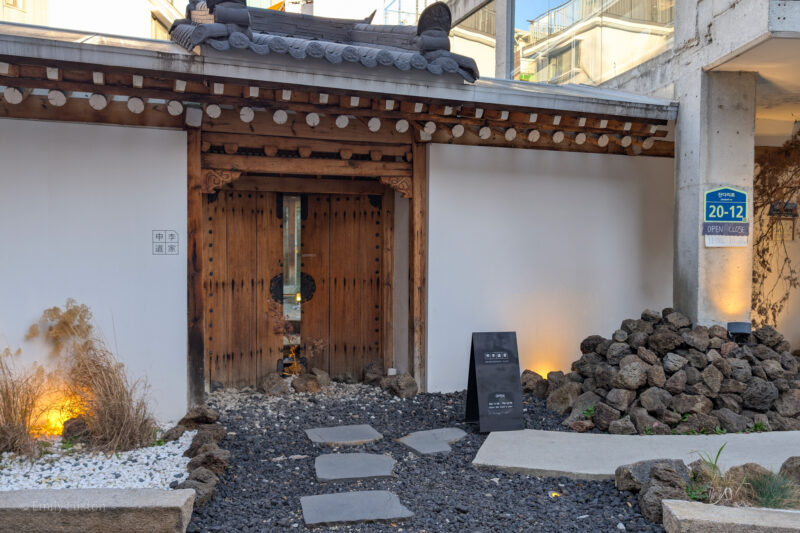 The entrance to a traditional Korean building with a wooden gate and tiled roof. The gate is flanked by white walls, and there is a small garden area with stepping stones and rocks leading up to the entrance. A sign on the right side of the entrance shows the address "20-12" and the opening hours "OPEN 11:00 - CLOSE 18:00."