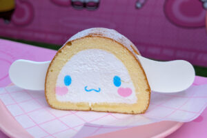 A slice of roll cake decorated to look like a cute character with blue eyes, pink cheeks, and a small blue mouth. The cake has white cream filling and is dusted with powdered sugar.