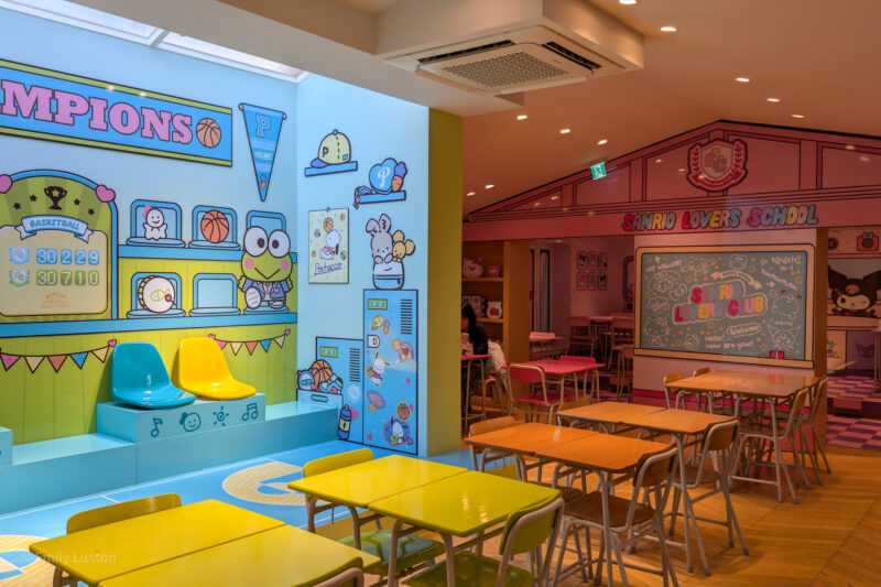 A colorful, cartoon-themed indoor space with Sanrio decorations, including a section labeled "CHAMPIONS" with a basketball theme and a scoreboard, and a "SANRIO LOVERS SCHOOL" classroom-like setting with tables, chairs, and Sanrio characters.