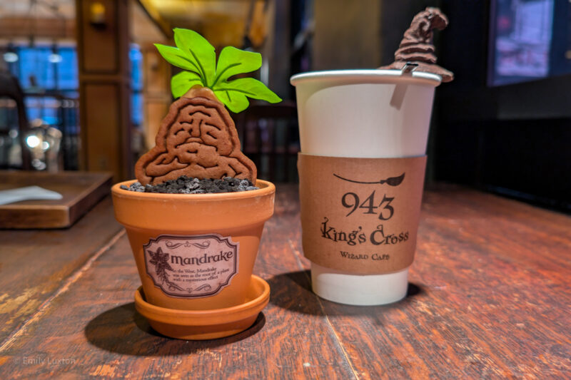 Two items on a wooden table: a terracotta pot with a mandrake root figure, labeled "mandrake," and a white paper cup with a "943 King's Cross Wizard Café" sleeve, topped with a chocolate swirl resembling a wizard's hat.