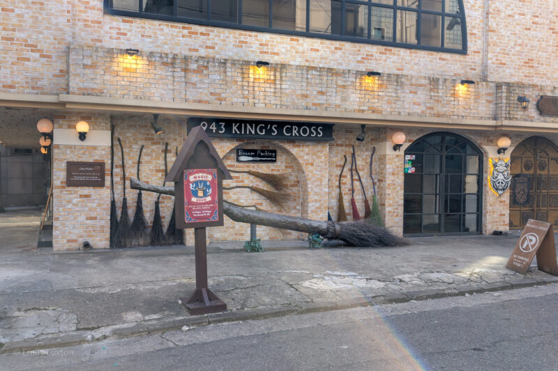 The exterior of the Harry Potter themed cafe in Seoul, with a sign that reads "943 KING'S CROSS." The building has a brick facade with several broomsticks leaning against the wall,. A signpost in the foreground advertises a "Magic Show" and indicates "Broom Parking." The overall theme is inspired by the Harry Potter series, referencing King's Cross Station and magical elements like broomsticks.