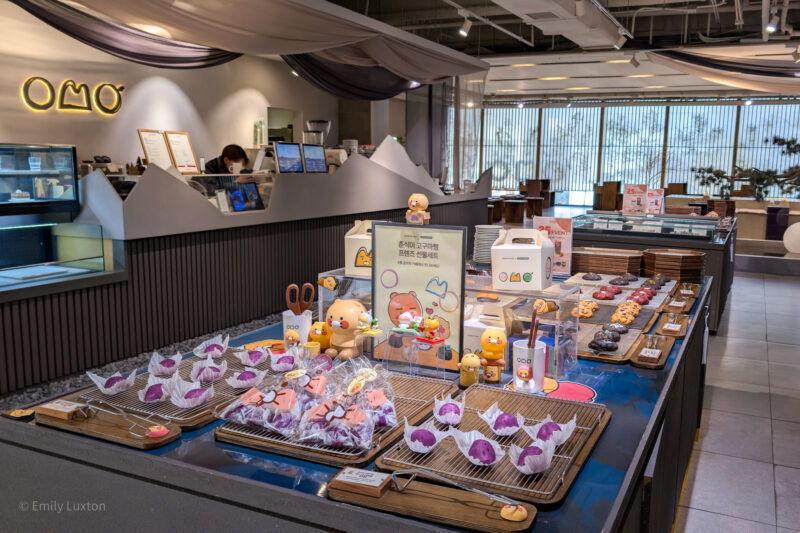 The interior of a café with a display table filled with various baked goods, including purple pastries and character-shaped cookies. The table is decorated with small figurines and a sign with Korean text. In the background, there is a counter with a neon sign that reads "OMO," and behind the counter, staff members are working. The setting is modern and well-lit, with large windows allowing natural light to enter.