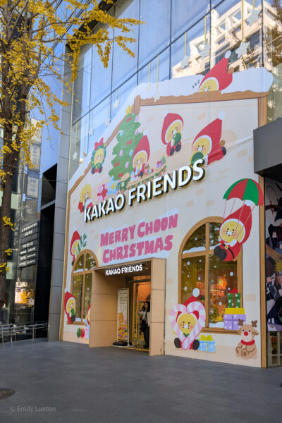 The exterior of a Kakao Friends store decorated for Christmas. The storefront features large, colorful illustrations of Kakao Friends characters dressed in festive attire, such as Santa hats and elf costumes. The decorations include a Christmas tree, presents, and various holiday-themed elements. The text "MERRY CHOON CHRISTMAS" is prominently displayed above the entrance. The store's name, "KAKAO FRIENDS," is also visible in large letters above the entrance. The surrounding area includes a tree with yellow leaves and a modern building facade.