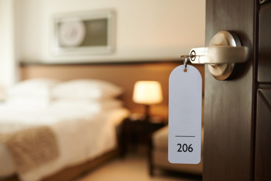 Hotel room with a slightly open door. A key tag with the number "206" is attached to the door handle. The background shows a neatly made bed, bedside table with a lamp, and a framed picture on the wall.