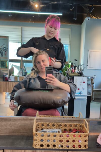 Emily sits in a salon chair, having her hair styled by a hairdresser. Emily is wearing a grey salon robe and holds a phone, capturing the moment, while the hairdresser works on their hair. The counter in front of them has a basket with various items, and salon equipment is visible in the background. The hair stylist is a Korean woman wearing a black top and has bright pink hair tied up in a ponytail.