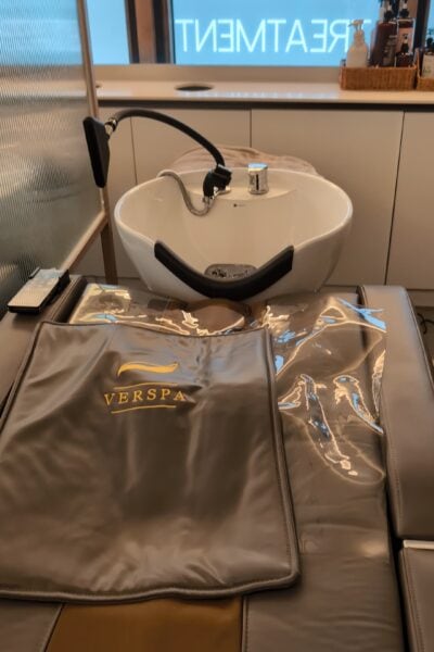 Salon or spa setting featuring a hair washing station with a white ceramic sink, black neck rest, and a padded chair covered in plastic. The background shows a counter with bottles, a basket, and a frosted glass partition with the word "TREATMENT" partially visible.