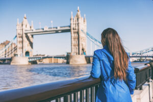 A person in a blue coat stands by a railing, gazing at the Tower Bridge in London on a clear day.