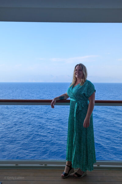 Emily standing on the deck of a ship, leaning against the railing. She is wearing a long, green dress with a floral pattern and black wedge sandals. The background features a vast expanse of blue ocean with a faint outline of distant mountains or land on the horizon. The sky is clear with a few scattered clouds.