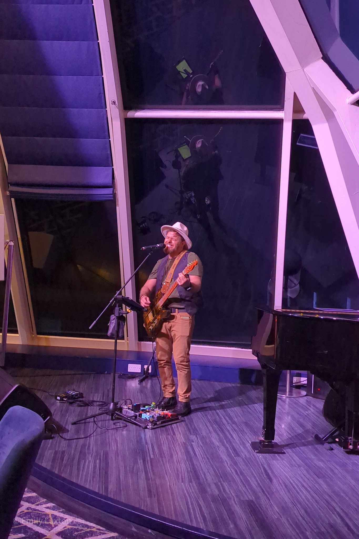 A musician is performing on stage, playing an electric guitar and singing into a microphone. The musician is wearing a hat, t-shirt, and pants. There is a music stand in front of the musician, and various guitar pedals are placed on the floor. The stage is set indoors with a large window behind the musician, reflecting the scene. A grand piano is also visible on the right side of the image,