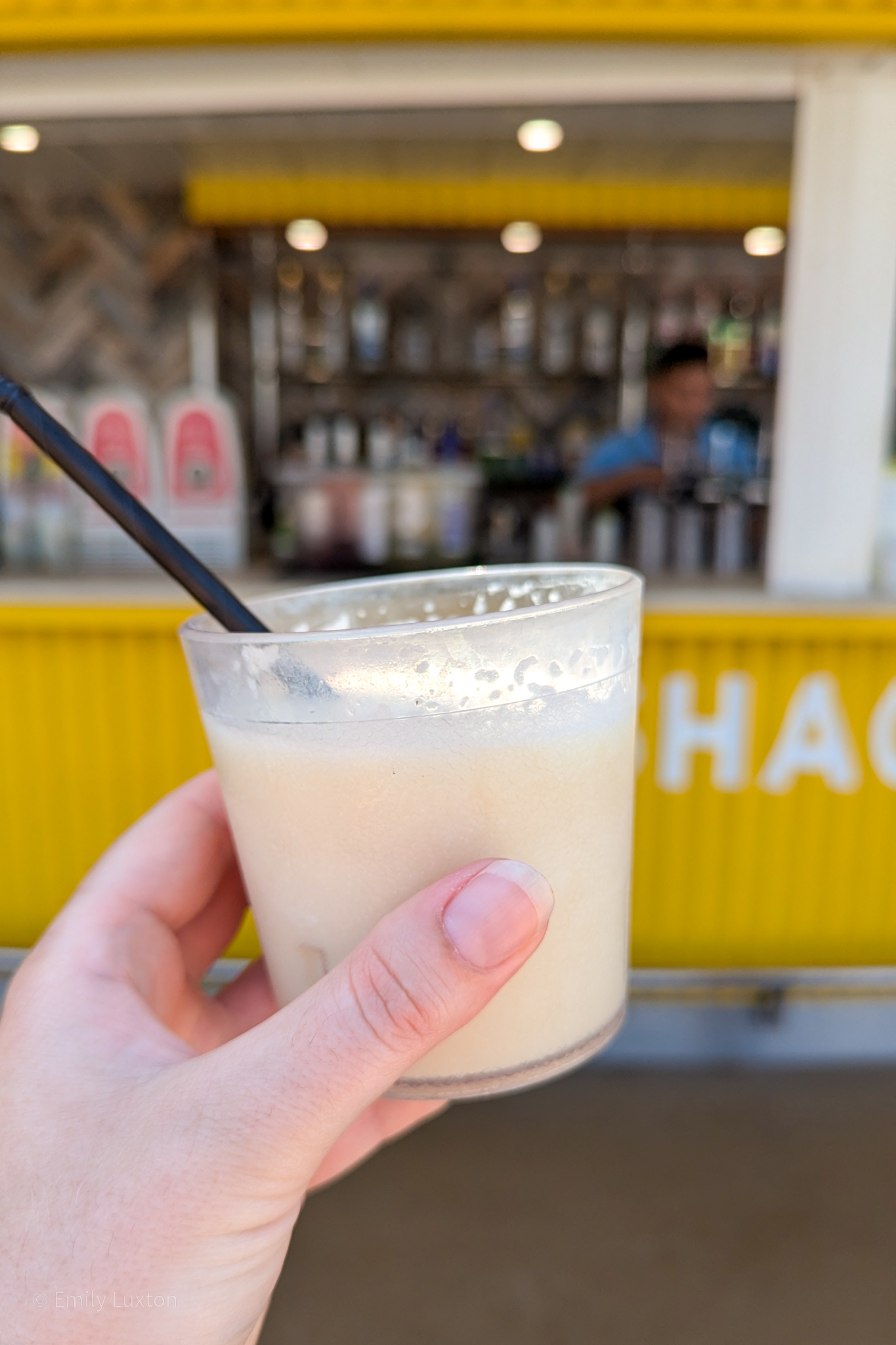 A hand holds a glass filled with a light-colored, frothy beverage with a black straw in it. The background shows a yellow structure with the partially visible word "SHACK". The image also includes a bar area with various bottles and dispensers.